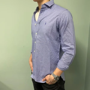 Blå skjorta från Ralph Lauren - Snygg blå skjorta från Ralph Lauren med diskret rutmönster och klassisk krage. Modellen är 190cm. Hör av er vid intresse!! 