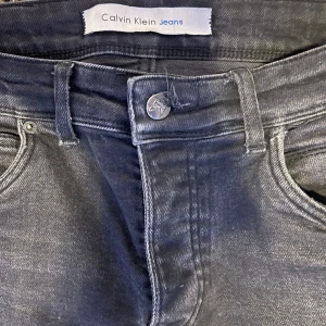 Svarta jeans från Calvin Klein - Snygga svarta jeans från Calvin Klein med klassisk femficksdesign och knappgylf. Perfekta för en stilren look. Passar till alla tillfällen!