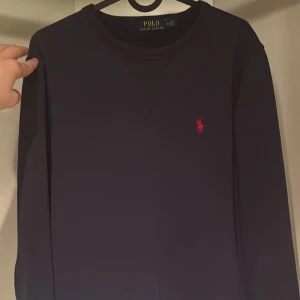 Mörkblå tröja från Polo Ralph Lauren - Säljer en stilren mörkblå tröja från Polo Ralph Lauren med det klassiska röda logotypbroderiet på bröstet. Tröjan har långa ärmar och en rund halsringning. Perfekt för en avslappnad och snygg look.