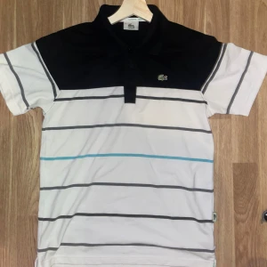 Randig pikétröja från Lacoste - Säljer en stilren pikétröja från Lacoste med svart krage och randigt mönster i svart och blått på vit bakgrund. 🐊🐊🐊