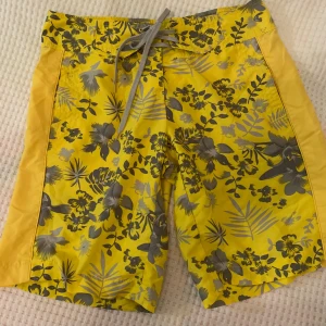 Gula blommiga shorts från Only - Snygga gula shorts från Only med ett grått blommönster. De har en bekväm passform med snörning i midjan för justerbarhet. Perfekta för en solig dag på stranden eller en avslappnad sommarutflykt.
