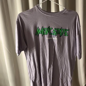Lila Magic State t-shirt - Säljer en lila t-shirt med tryck från Magic State. T-shirten har korta ärmar och ett coolt grafiskt tryck i grönt och vitt på framsidan och baksidan. Perfekt för dig som gillar unika bandtröjor!