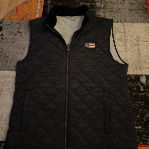 Mörk blå quiltad väst från Lexington - Säljer en stilren mörkblå quiltad väst från Lexington med dragkedja och en broderad logotyp på bröstet. Perfekt för lager-på-lager under kyligare dagar. Västen har en praktisk innerficka med dragkedja.