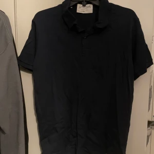 Mörkblå kortärmad pikétröja från Selected Homme - Snygg mörkblå kortärmad pikétröja från Selected Homme. Perfekt för en avslappnad stil med klassisk krage och knappstängning framtill. Skjortan är tillverkad i ett mjukt material som ger en bekväm passform.