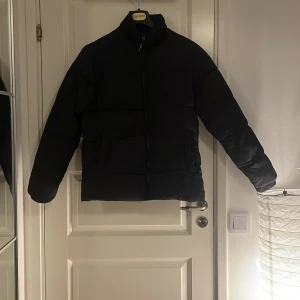 Svart pufferjacka från Jack & Jones - Säljer en svart pufferjacka från Jack & Jones i storlek M. Jackan har en stilren design med dragkedja och långa ärmar. Perfekt för kyligare dagar. Har ett litet hål bak på höger sidan av ryggen. Annars är den i bra skick👌🏻pris kan diskuteras⚜️