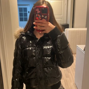 Svart dunjacka från Moncler - Säljer en snygg svart dunjacka från Moncler med glansig finish och pälskantad huva. Jackan har dragkedja och knappar framtill samt ribbade muddar. Perfekt för kyliga dagar! Finns inget äktighets bevis 