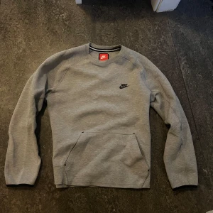 Grå tröja från Nike - Säljer en stilren grå tröja från Nike med lång ärm och en liten Nike-logga på bröstet. Tröjan har en praktisk ficka framtill och är perfekt för en avslappnad stil.