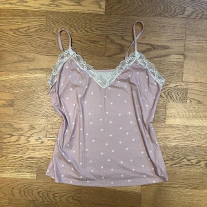 Ljusrosa prickig linne med spetsdetaljer - Säljer en söt ljusrosa linne med vita prickar och spetsdetaljer vid halsringningen. Linnet har smala axelband och en v-ringad design. Perfekt för varma dagar eller som ett lager under en tröja.