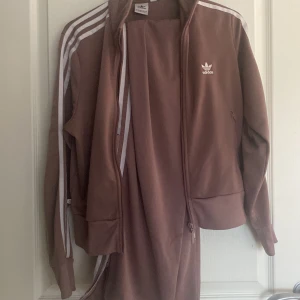  Mörk lika/risa träningset från Adidas strl L - Säljer en brun träningsjacka och matchande byxor från Adidas. Jackan har dragkedja och de klassiska tre ränderna på ärmarna. Byxorna har elastisk midja och ränder längs sidorna. Perfekt för en sportig look! Båda för 175kr annars 100kr st