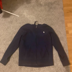 Mörkblå hoodie från Ralph Lauren - Säljer en stilren mörkblå hoodie från Ralph Lauren med dragkedja och det klassiska broderade logotypen på bröstet. Perfekt för en avslappnad och snygg look. Passar bra till både jeans och chinos.