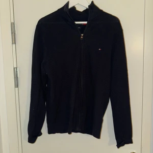 Svart stickad tröja från Tommy Hilfiger - Säljer en stilren svart stickad tröja från Tommy Hilfiger. Tröjan har en dragkedja framtill och ett diskret broderat logotypmärke på bröstet. Perfekt för kyligare dagar.