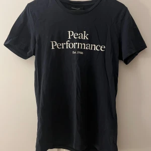 Svart t-shirt, Peak Performance - Snygg svart t-shirt från Peak Performance med vit text på framsidan. Perfekt för en stilren och avslappnad look. Tillverkad i mjukt material för extra komfort.