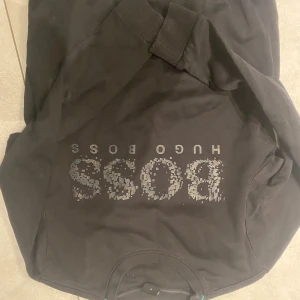 Svart tröja från Hugo Boss - Säljer en stilren svart tröja från Hugo Boss med deras logga i ett coolt mönster på framsidan. Tröjan har långa ärmar och ribbade muddar vid ärmslut och nederkant. Perfekt för en casual look.