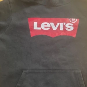 Svart hoodie från Levi's - Säljer en svart hoodie från Levi's med det klassiska röda och vita logotyptrycket på bröstet. Tröjan har en stor ficka framtill och en bekväm huva. Perfekt för en avslappnad stil.