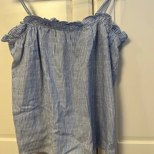 Blå- och vitrandig linnetopp - Säljer en söt blå- och vitrandig linnetopp med smala axelband och volangdetaljer vid halsringningen. Perfekt för varma sommardagar. Toppen har en luftig och bekväm passform.