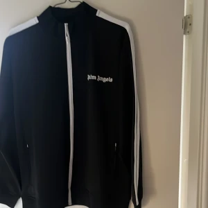 Svart track jacket från Palm Angels - Säljer en svart track jacket från Palm Angels med vita detaljer. Tröjan har en dragkedja framtill och vita ränder längs ärmarna. Den har en hög krage och Palm Angels-loggan på bröstet. Perfekt för en sportig look! Aldrig använd då den är för stor för mig. Storlek L men skulle eventuellt kunna passa M