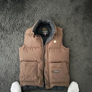 Brun dunväst från Canada Goose - Säljer en brun dunväst från Canada Goose med en stilren design. Västen har en hög krage och knappar framtill. Den är perfekt för kyligare dagar och har praktiska fickor för förvaring. Riktigt fint skick inte alls använd, och så är den ju såklart äkta! Det är storlek xs men skulle säga att den passar S också!