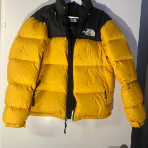 Gul pufferjacka från The North Face - Säljer en cool gul pufferjacka från The North Face med svarta detaljer. Det finns en lagning på vänster arm, men jäkligt skickligt gjort så den flyter in fint i jackan. 