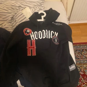 Svart hoodie från Hoodrich - Säljer en svart hoodie från Hoodrich med flera coola patches och broderade detaljer. Den har en stor H-logga och texten 'Hoodrich' på framsidan. Perfekt för en avslappnad stil. Den är i bra skick skriv om ni har några frågor 