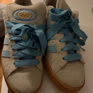 Adidas Campus sneakers med blå detaljer - Snygga Adidas Campus sneakers i beige mocka med ljusblå snörning och detaljer. Skorna har en klassisk design med gummisula och det ikoniska tre-randiga mönstret på sidan. Perfekta för en stilren och avslappnad look.