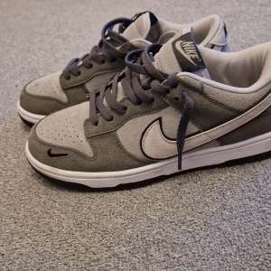 Grå Nike Dunk sneakers - Snygga grå Nike Dunk sneakers med vit sula och klassisk Nike-logga på sidan. Skorna har snörning och är tillverkade i mocka och syntetmaterial. Perfekta för en stilren look.