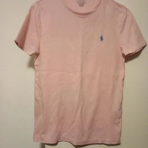 Rosa t-shirt från Ralph Lauren - Säljer en stilren rosa t-shirt från Ralph Lauren med en liten blå broderad logga på bröstet. T-shirten har en custom slim fit passform och är tillverkad i mjuk bomull. Perfekt för en avslappnad look.Ny T-shirt från Polo, aldrig använt råkade få fläckar under tvätt se bilderna fläkar är inte en stor grej. Den är helt nytt. Ord. pris: 899kr