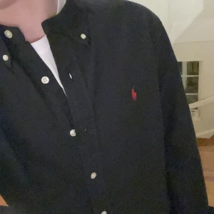 Svart skjorta från Ralph Lauren - Säljer en stilren svart skjorta från Ralph Lauren med klassisk krage och knappar. Den har en broderad röd logga på bröstet och är långärmad. Perfekt för en snygg och avslappnad look.