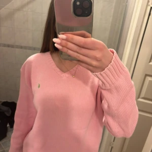 Rosa stickad tröja från Polo Ralph Lauren - Säljer en snygg rosa stickad tröja från Polo Ralph Lauren med det klassiska gröna logotypen på bröstet. Tröjan har en v-ringad hals och långa ärmar, perfekt för en stilren look. Passar bra till både jeans och kjol. 🌸Finns ställen där trådar sticker ut, skriv för fler bilder! 
