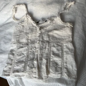 Vit ärmlös blus med spetsdetaljer - Charmig vit blus med knappar framtill och spetsdetaljer. Den är ärmlös med tunna axelband och har en luftig design. Perfekt för varma dagar. Har dessvärre defekter som syns i bilderna.