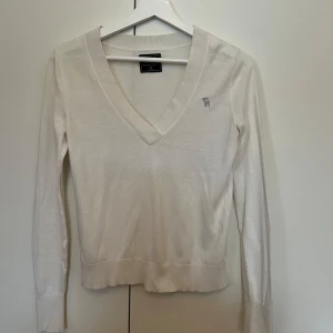 Tröja från Abercrombie & Fitch - Säljer en stilren vit v-ringad tröja från Abercrombie & Fitch. Tröjan har långa ärmar och en liten broderad logotyp på bröstet. Perfekt för en avslappnad och snygg look.