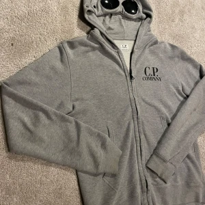 Grå huvtröja från C.P. Company - Säljer en grå huvtröja från C.P. Company med dragkedja och unika glasögondetaljer i huvan. Perfekt för en stilren och avslappnad look. Tröjan har långa ärmar och ribbade muddar för extra komfort. Nypris: 2200kr, Mitt pris: 659kr