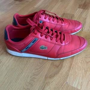 Sneakers från Lacoste - Snygga röda sneakers från Lacoste med klassisk snörning och vit sula. Skorna har Lacoste-loggan på sidan och en mörkblå detalj vid hälen. Perfekta för en sportig look. Kontakta gärna vid fler frågor 😃Nypris: 1200kr 