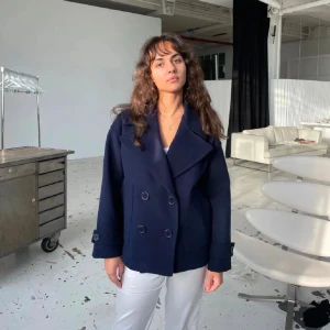 Mörkblå kappa från Design by Si - Snygg mörkblå kappa från Design by Si med dubbelknäppning och bred krage. Jackan har långa ärmar och en stilren design som passar perfekt för kyligare dagar. Perfekt för dig som vill ha en klassisk look.