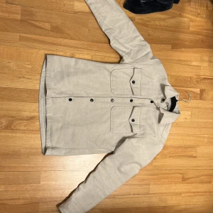 Vit overshirt med svarta knappar - Säljer en stilren vit overshirt med knappar framtill och två bröstfickor. Perfekt för lager-på-lager-stil och passar till många olika outfits. Den har långa ärmar och en klassisk krage. Xs