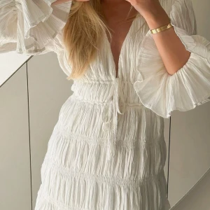 Vit studentklänning med volangdetaljer - Säljer en söt vit klänning med volangdetaljer och knytning framtill. Klänningen har långa ärmar och en v-ringad design som ger en romantisk känsla. Perfekt för sommarens alla tillfällen! Perfekt studentklänning med lappen kvar!! SLUTSÅLD🥰🥰