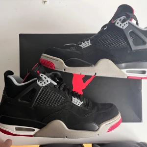 Nike Air Jordans 4 bred - Snygga Nike Air Jordans 4 breds i svart med röda detaljer. Skorna har en klassisk design med snörning och en robust sula. Perfekta för dig som vill ha en stilren look med en touch av färg.
