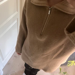 Beige stickad tröja med dragkedja - Säljer en supermysig beige stickad tröja med dragkedja framtill. Perfekt för kyliga dagar med sin långa passform och ribbade struktur. Tröjan har långa ärmar och en bekväm krage som kan justeras med dragkedjan. Passar perfekt till både vardag och mysiga hemmakvällar.
