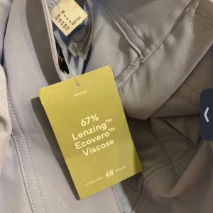 Grå kjol från H&M - Säljer en snygg blå kjol från H&M i storlek 38. Kjolen är tillverkad av 67% Lenzing Ecovero Viscose, vilket gör den både bekväm och miljövänlig. Den har ett stilrent utseende med veckade detaljer och två vita ränder längst ner. Aldrig använd 