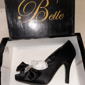 Säljer ett par eleganta svarta pumps från Belle. Skorna har en spetsig tå och en vacker rosett med pärldetalj framtill. De är tillverkade i ett glansigt material som ger en lyxig känsla. Perfekta för festliga tillfällen!