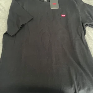Svart t-shirt från Levi's - Säljer en klassisk svart t-shirt från Levi's i storlek M. Den är gjord av mjuk bomull och har en liten röd Levi's-logga på bröstet. Perfekt för vardagsbruk och kan enkelt matchas med jeans eller shorts. T-shirten är i nyskick och har korta ärmar.