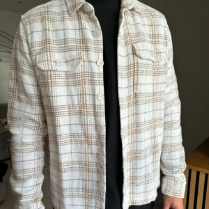 Rutig overshirt från New Look - Säljer en snygg rutig overshirt från New Look i storlek L. Den är i en beige och vit färgkombination med knappar framtill och två bröstfickor. Perfekt för höst och vår när du vill ha en stilren look.