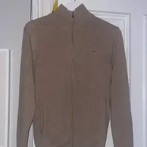 En stilren GANT zip-hoodie i en beige-brun färg, gjort ut av 100% lammull. Aldrig använd! Köpte den på El Corte Ingles i Palma för 1700