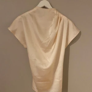 Champagnefärgad sidenklänning - Säljer en elegant champagnefärgad blus i siden. Den har en enkel och stilren design med korta ärmar. Perfekt för en festlig kväll eller en speciell tillställning.