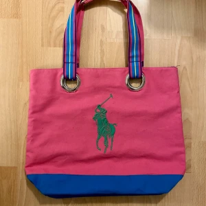 Ralph Lauren. Rosa och blå shoppingväska med polomotiv - Superfin rosa och blå shoppingväska från Ralph Lauren i bomullstyg. Väskan har ett grönt polomotiv på framsidan och färgglada randiga handtag. Perfekt för en dag på stan, till gymmet eller stranden! 🛍️ Hämtas på Hägerstensåsen eller ök. 