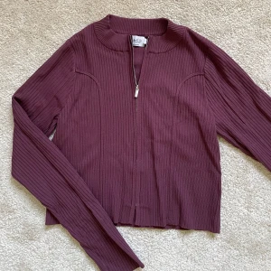 Vinröd ribbad kofta / cardigan L-M oanvänd  - Snygg vinröd kofta med ribbad textur och dragkedja framtill. Jackan har långa ärmar och en stilren design som passar perfekt för höst och vår. Perfekt för att ge din outfit en trendig touch!