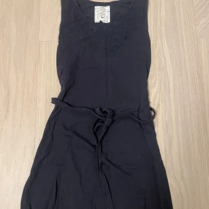 Svart ärmlös klänning från Emma och Malena - Snygg svart ärmlös klänning från Emma och Malena med broderade detaljer vid halsen. Klänningen har ett knytband i midjan för en justerbar passform. Perfekt för en stilren look.