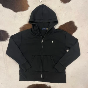 Svart hoodie från Polo Ralph Lauren - Säljer en stilren svart hoodie från Polo Ralph Lauren med dragkedja och det klassiska logotypen på bröstet. Perfekt för en avslappnad look. Den har långa ärmar och en praktisk huva.