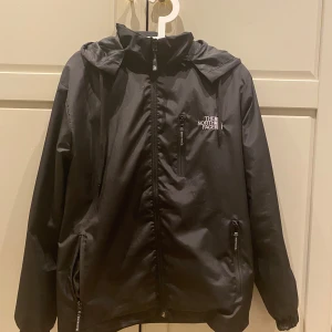 Svart vindjacka från The North Face - Säljer en svart vindjacka från The North Face med dragkedja och huva. Jackan har två sidofickor med dragkedjor och en liten logga på bröstet. Perfekt för blåsiga dagar. Inga defekter, pris går att diskutera.