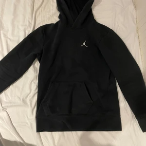 Svart hoodie från Jordan - Säljer en stilren svart hoodie från Jordan med den ikoniska loggan på bröstet. Tröjan har en praktisk magficka och en bekväm huva. Perfekt för en avslappnad look.