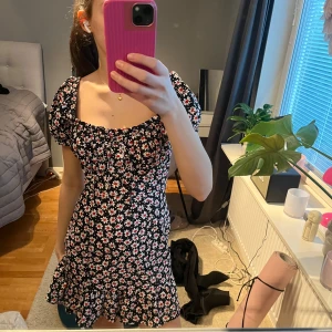 Blommig kortklänning med volangdetaljer - Söt kortklänning med blommigt mönster i svart, rosa och vitt. Lite insydd i midjan. Inte så använd 